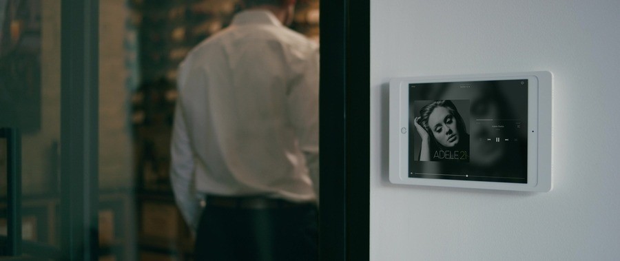 A wall-mounted tablet displaying Adele's music is used to control a multi-room audio system in a luxury home. A person stands in the background near a wine cellar. 